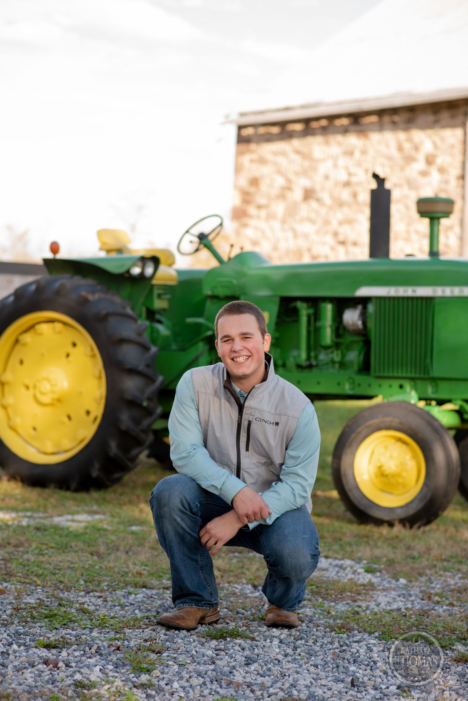 Person crouches beside classic John Deere tractor with stone barn in background.
