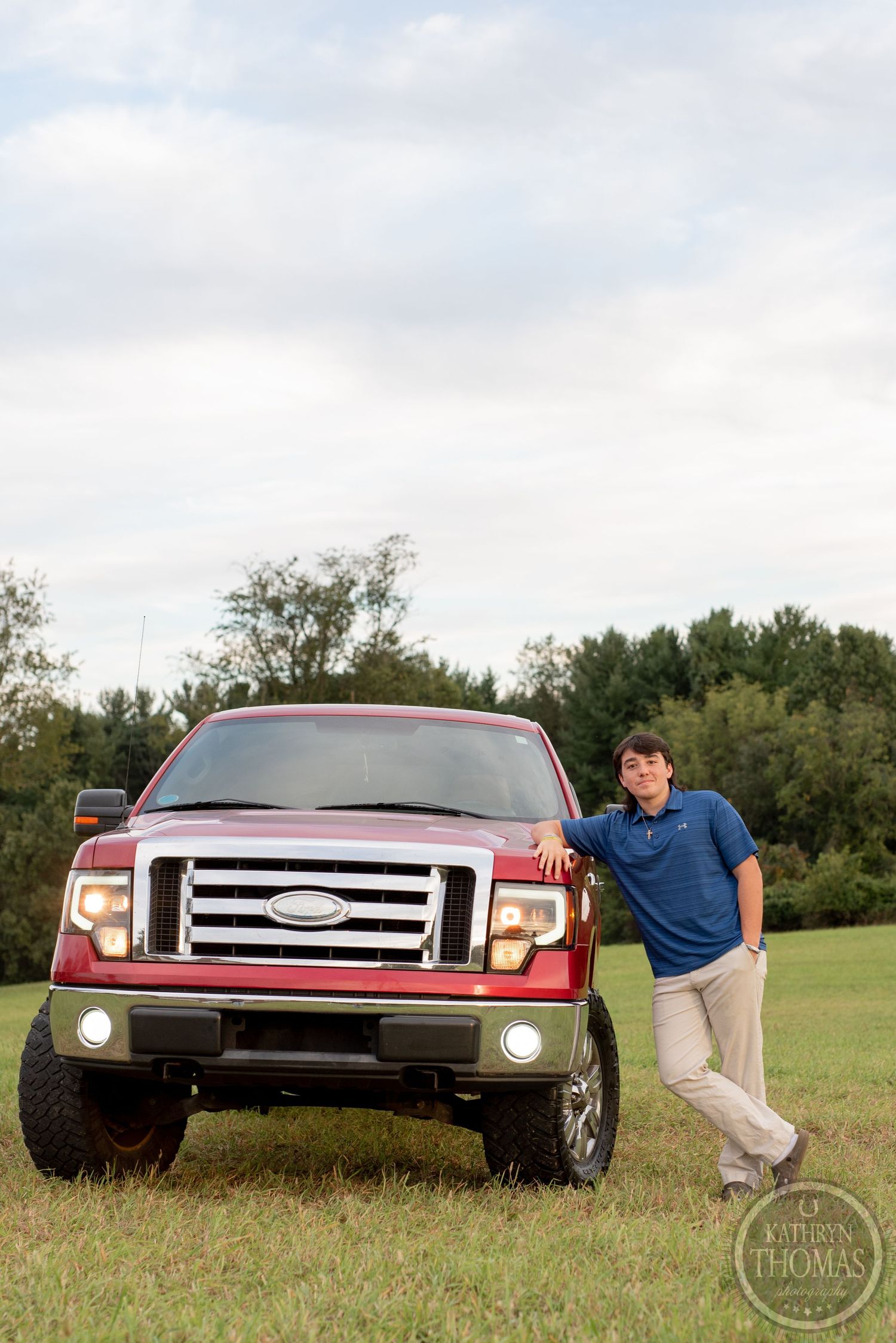 A person leans against a red Ford F150 with custom wheels parked in a grassy field.