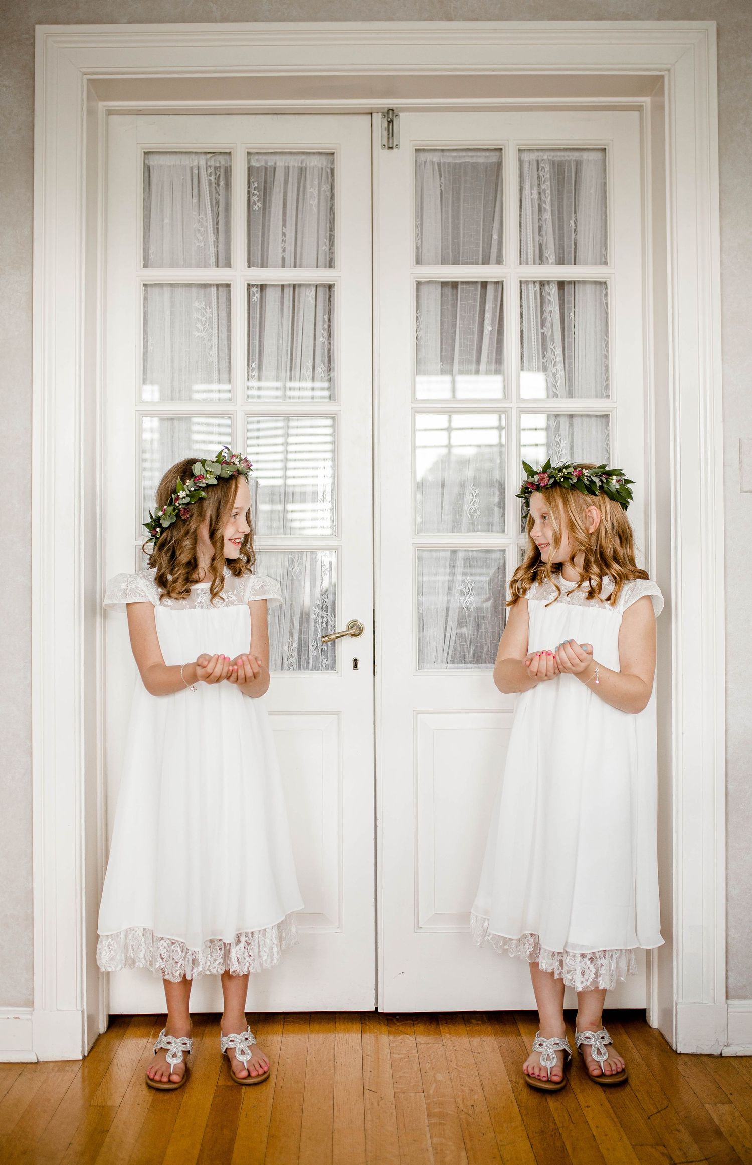 Two flower girls in white dresses and floral crowns stand symmetrically at French doors.