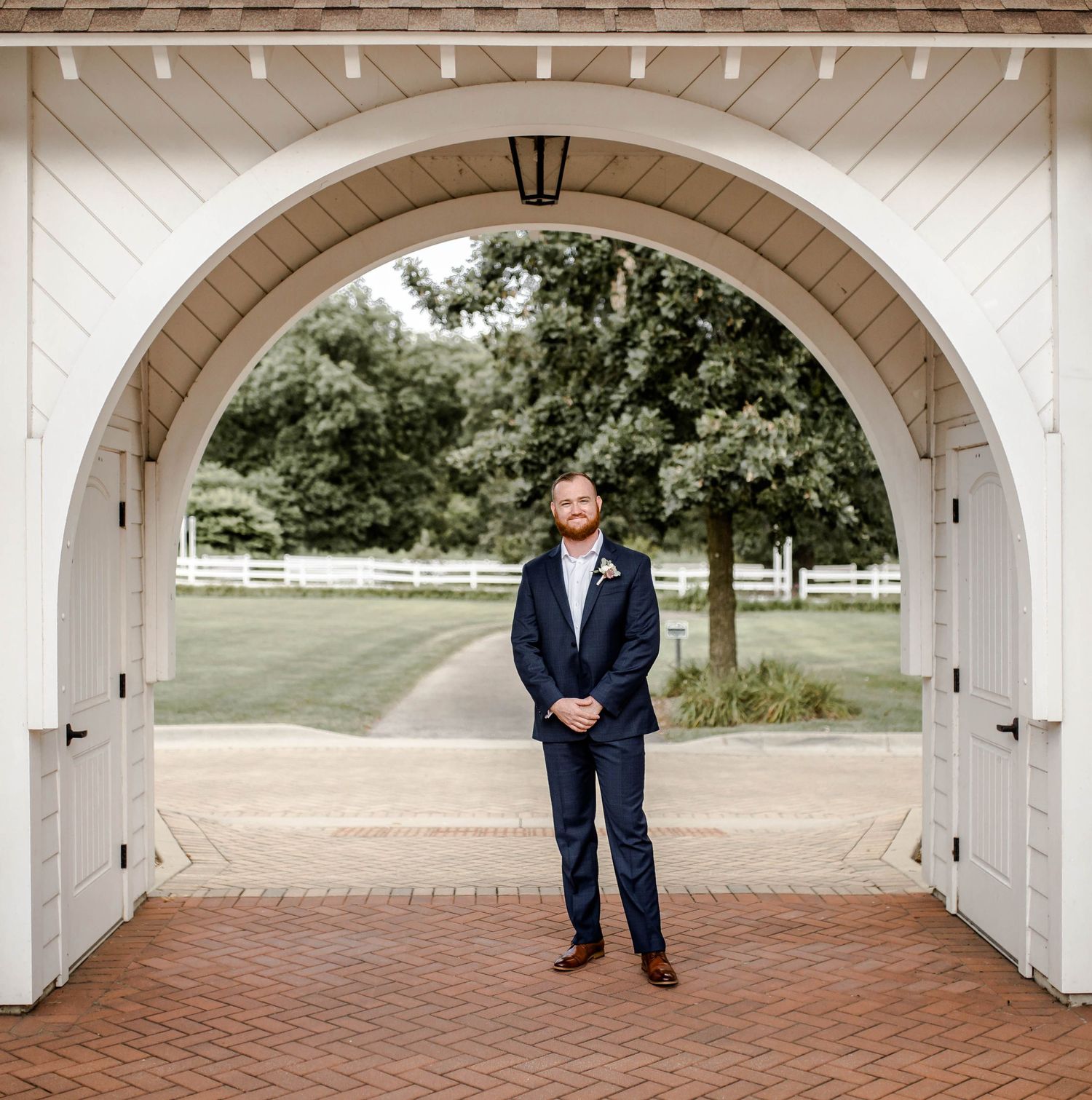 A figure stands under a white arched doorway framed by brick pavement and trees in the background.