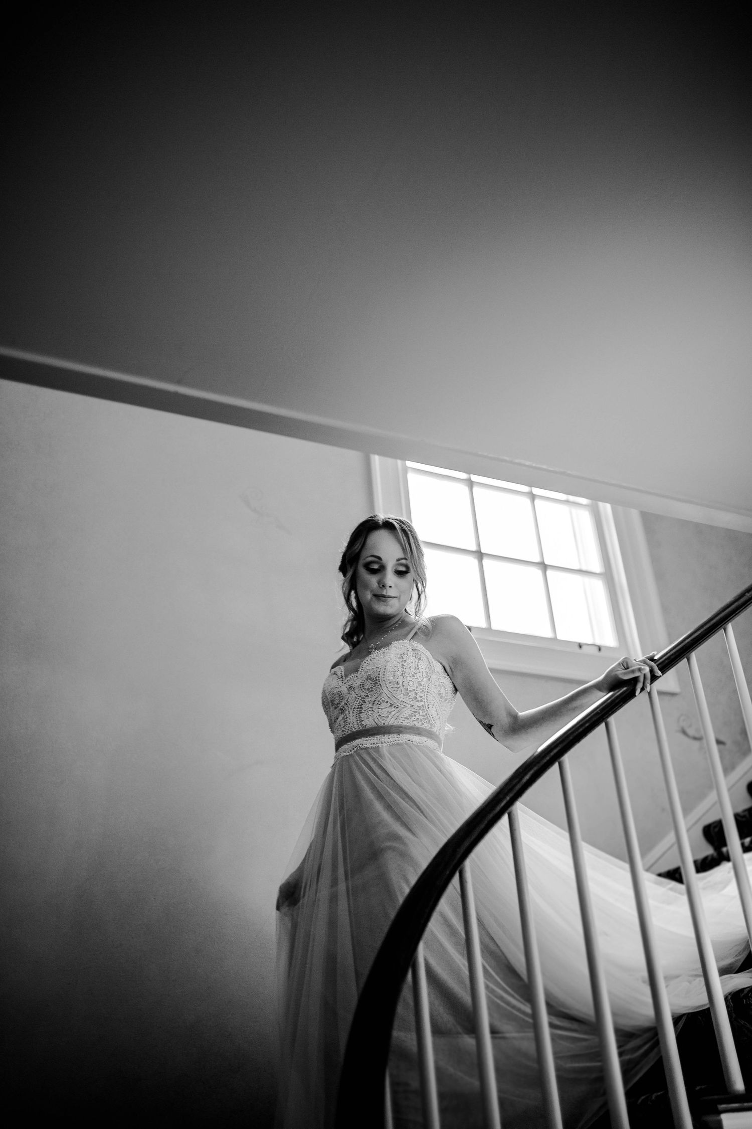 A bride in a flowing gown poses on an elegant curved staircase in black and white.