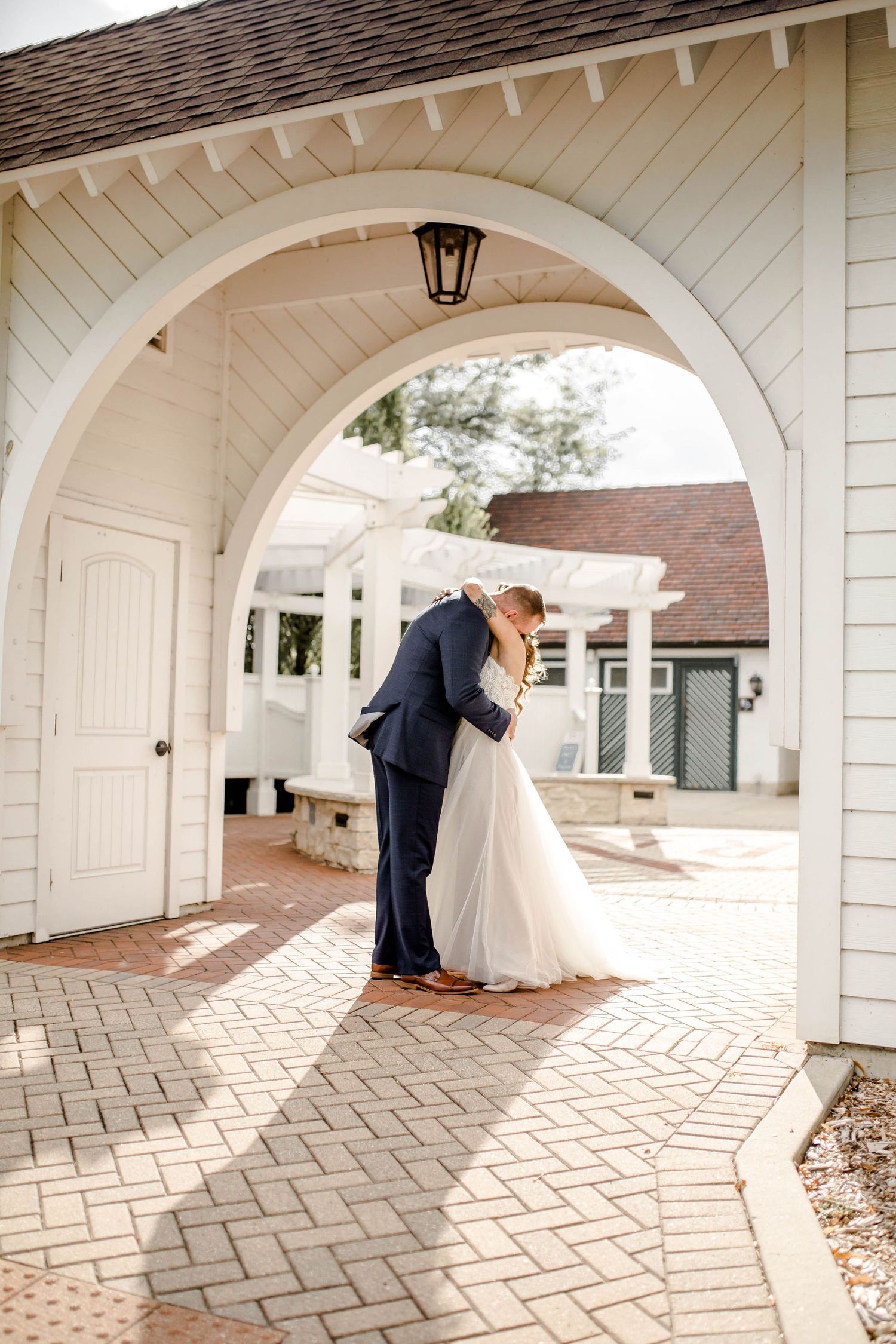 A wedding couple shares an intimate moment under a white architectural archway with brick pavement.