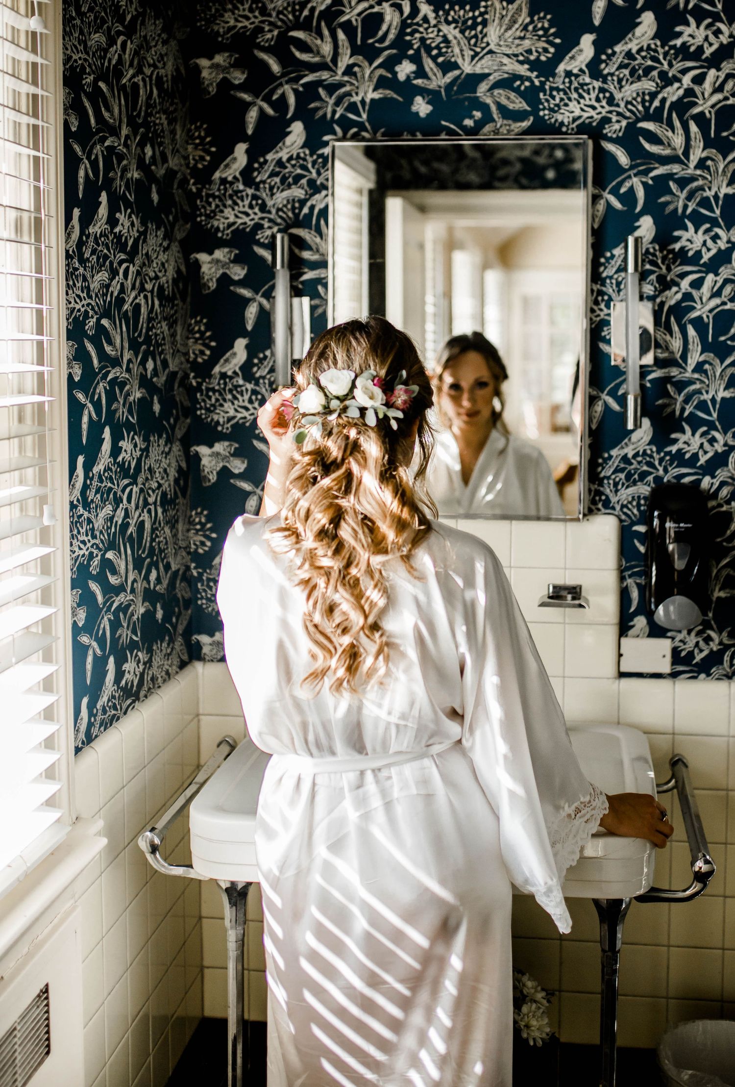 A white silk robe is reflected in a bathroom mirror with dramatic blue floral wallpaper.