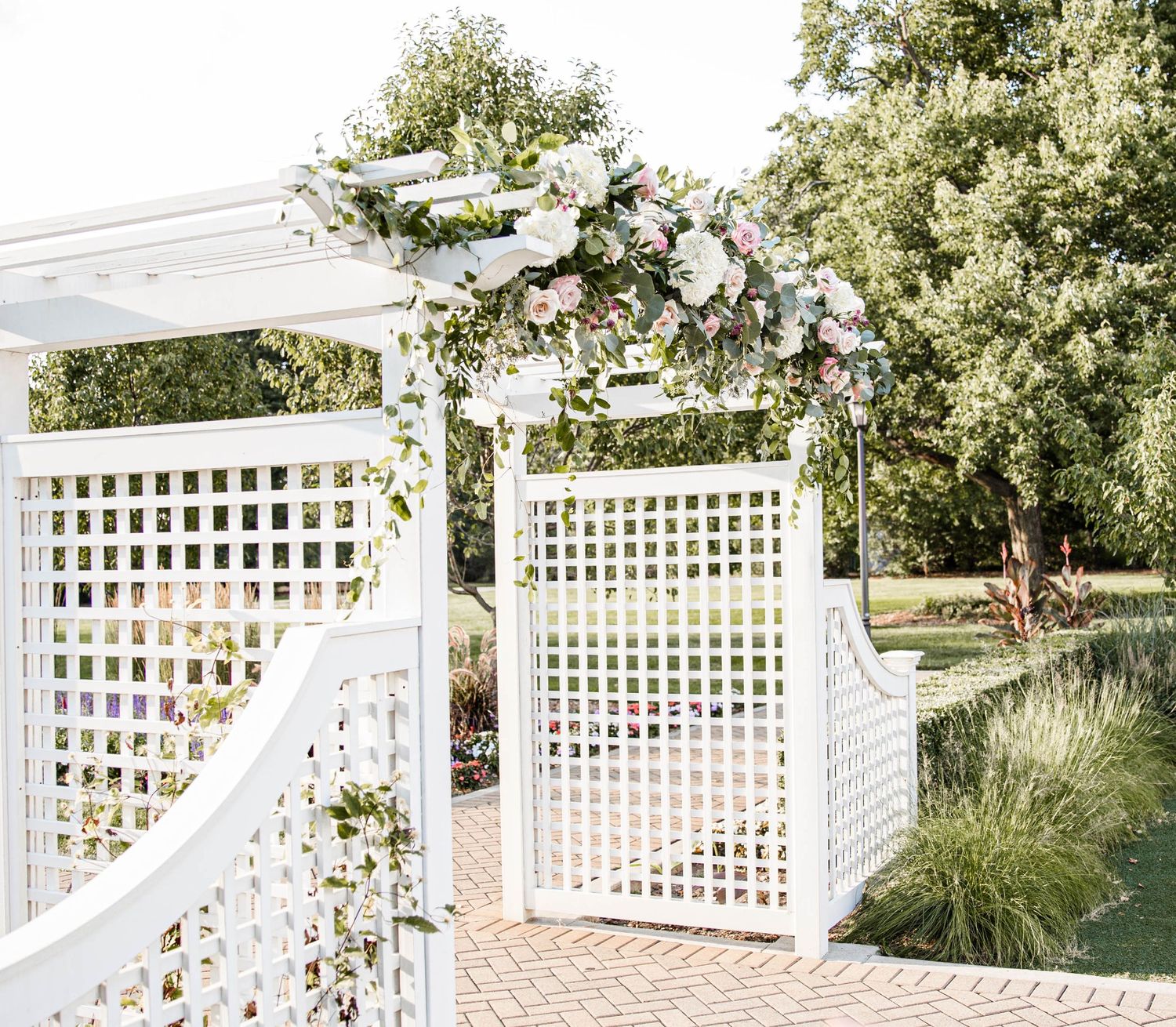 White lattice wedding arch decorated with floral arrangements in garden setting.