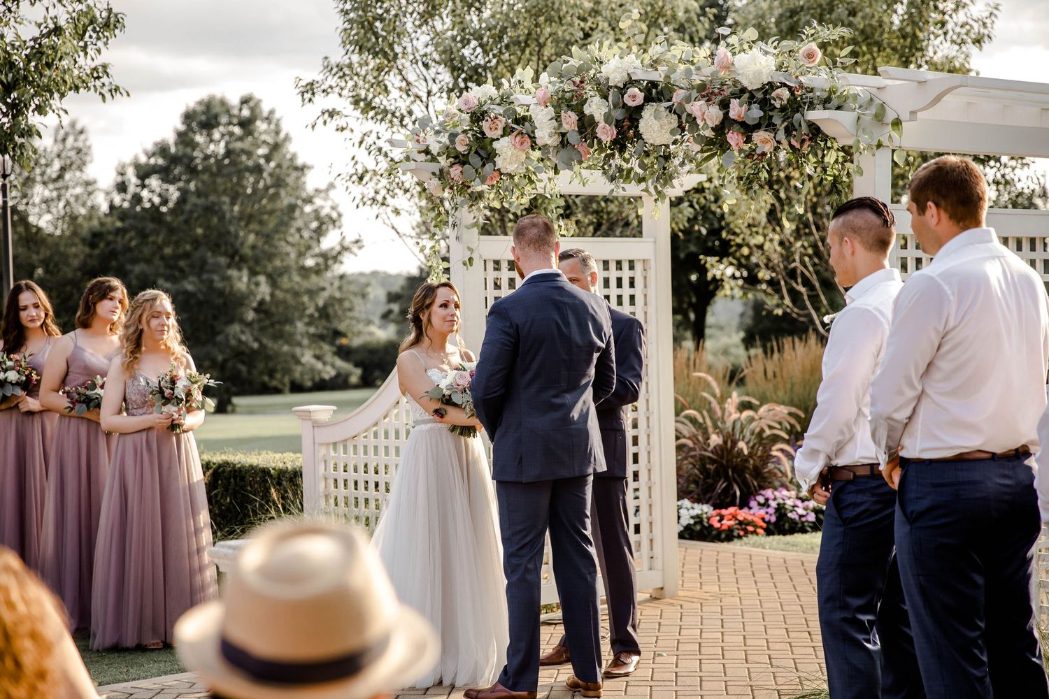An outdoor wedding ceremony takes place under a floral arch with attendees in purple dresses and formal attire.