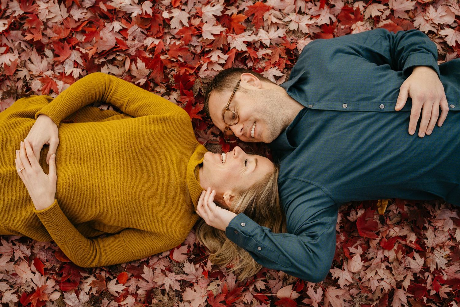 Couple lying on red autumn leaves wearing mustard yellow and navy blue sweaters in romantic outdoor setting.