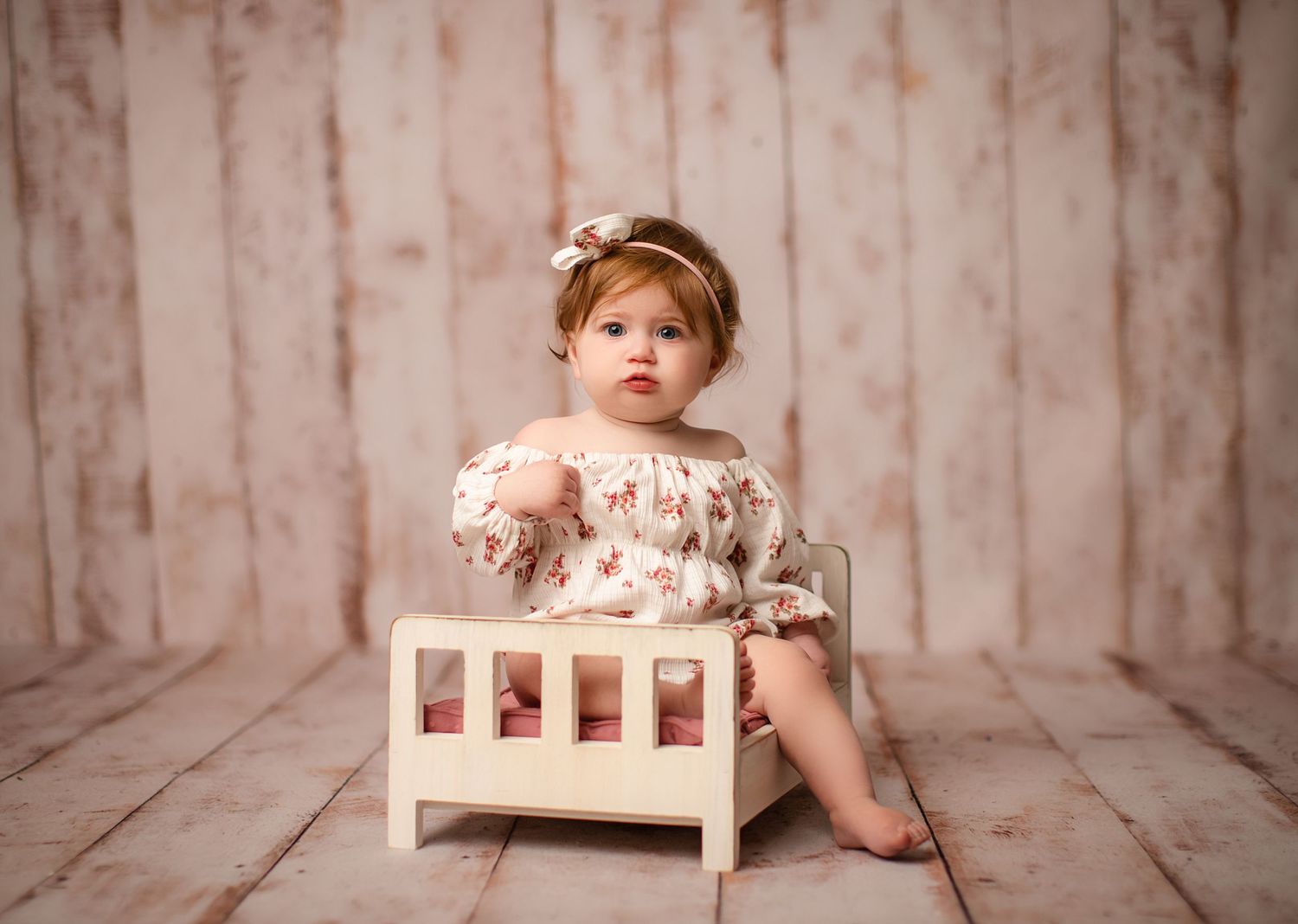Little one in floral dress sitting in small white wooden bed against rustic backdrop.
