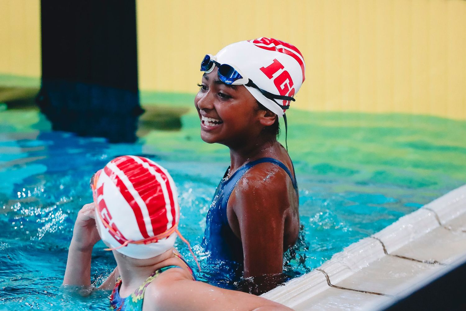 Two swimmers in red and white swim caps resting at pool edge during training.