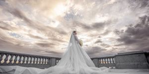 Stunning bride in flowing white gown with cathedral train stands on balcony under dramatic sunset sky.