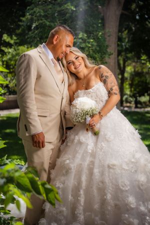 Bride in white floral ballgown holds white bouquet beside groom in beige suit in garden.