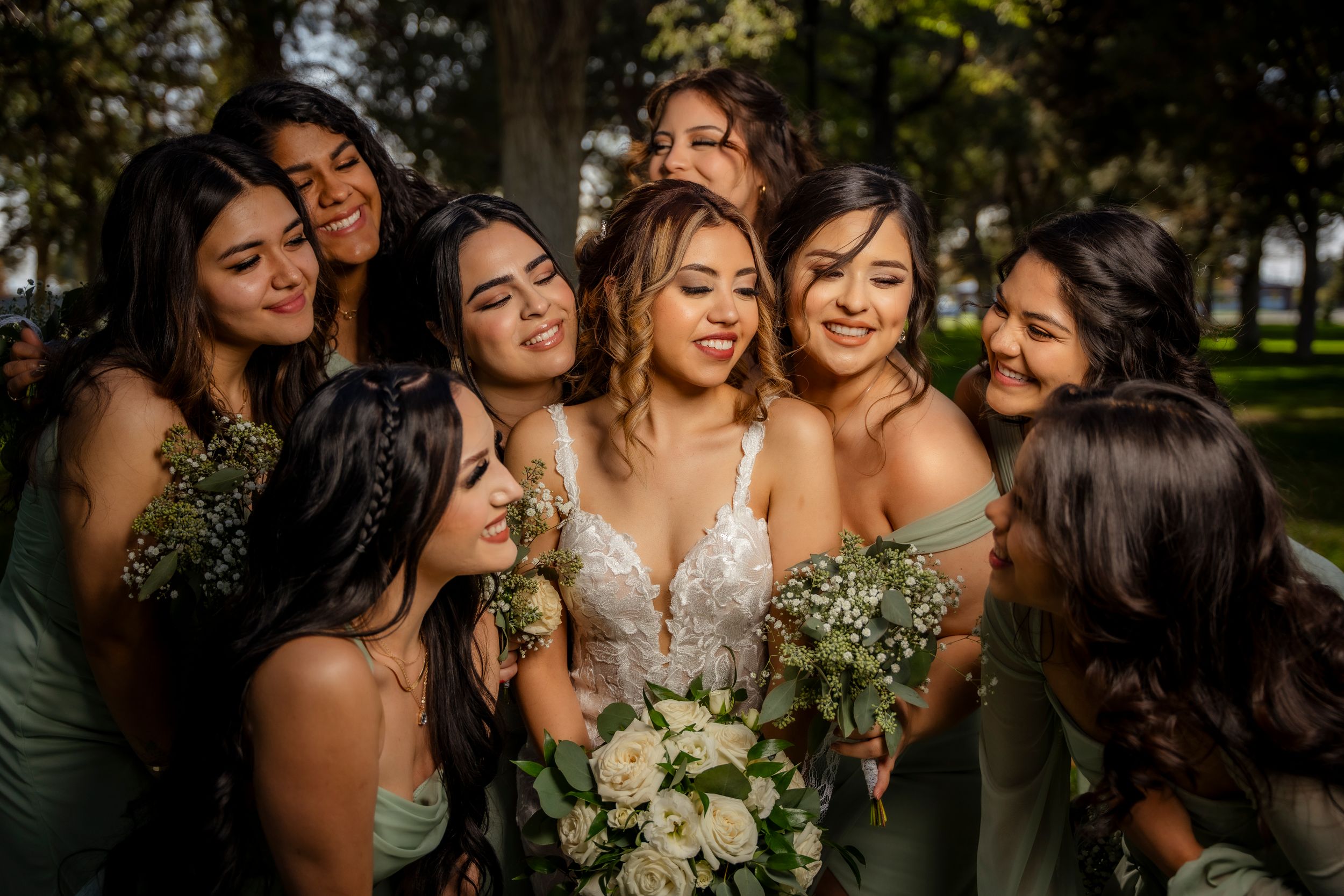 Bride in lace gown embraced by smiling bridesmaids in sage green dresses holding white bouquets outdoors.