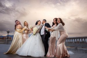 Bride and groom celebrate with four bridesmaids in gold dresses on rooftop under dramatic cloudy sky.