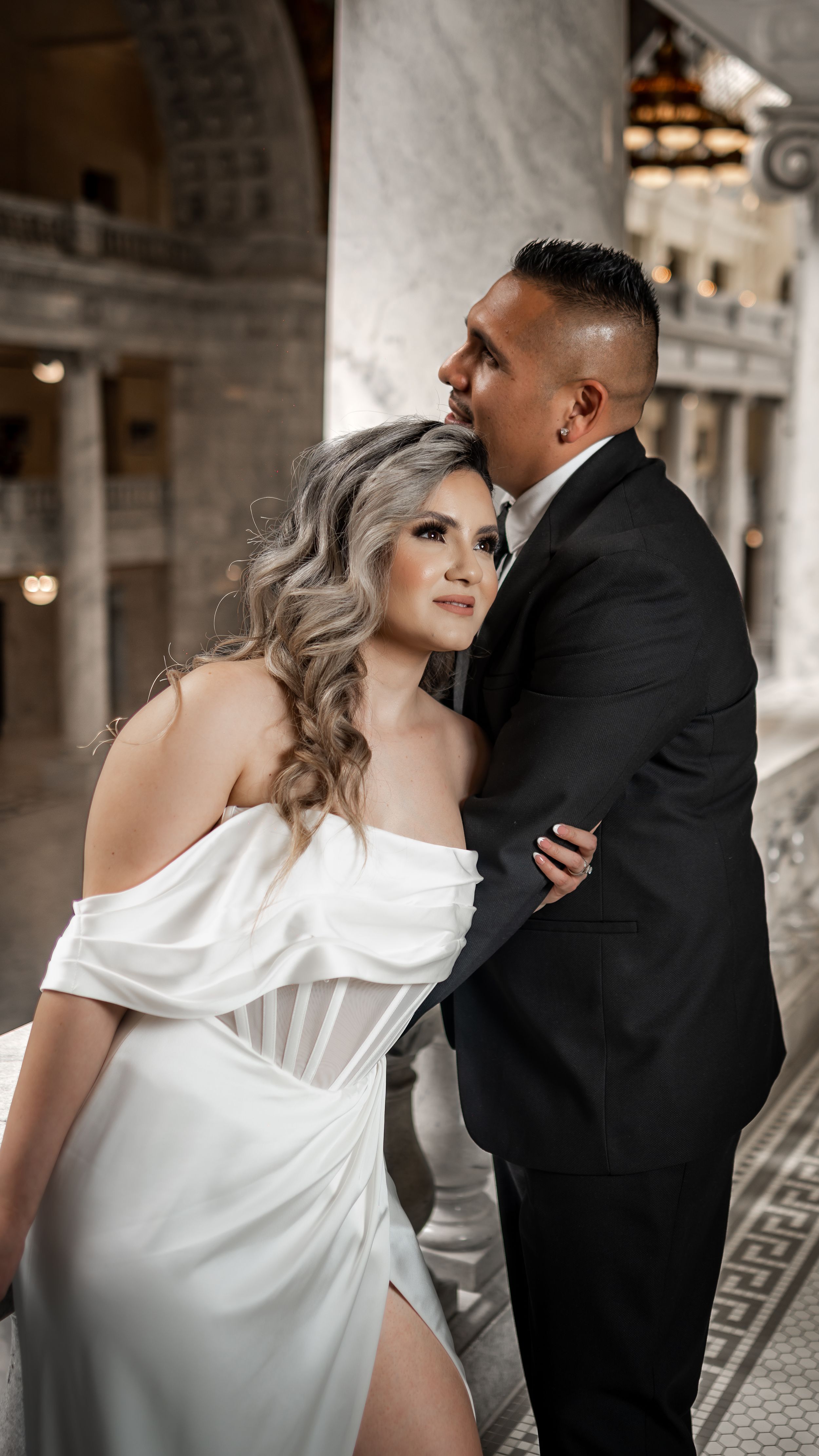 Elegant couple in white off-shoulder gown and black suit pose in marble architectural interior.