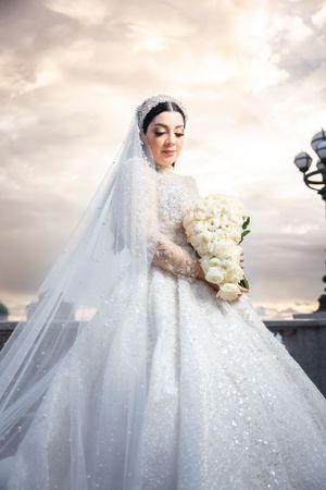 Elegant bride in sparkling white ball gown with long veil holds white floral bouquet under cloudy sky.