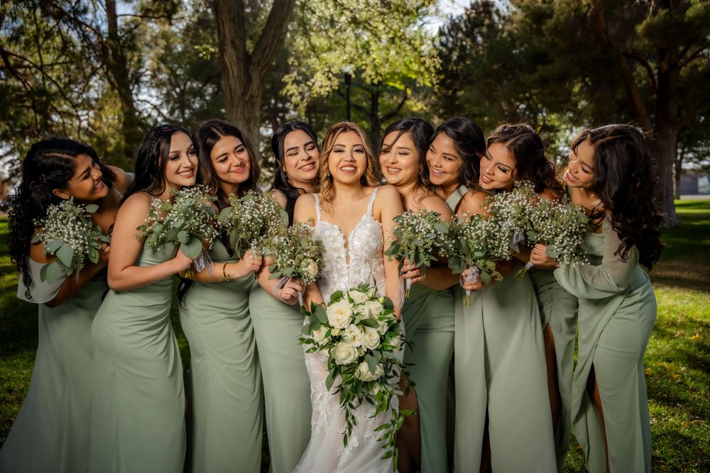 Bride surrounded by bridesmaids in sage green dresses holding white floral bouquets in sunny park.