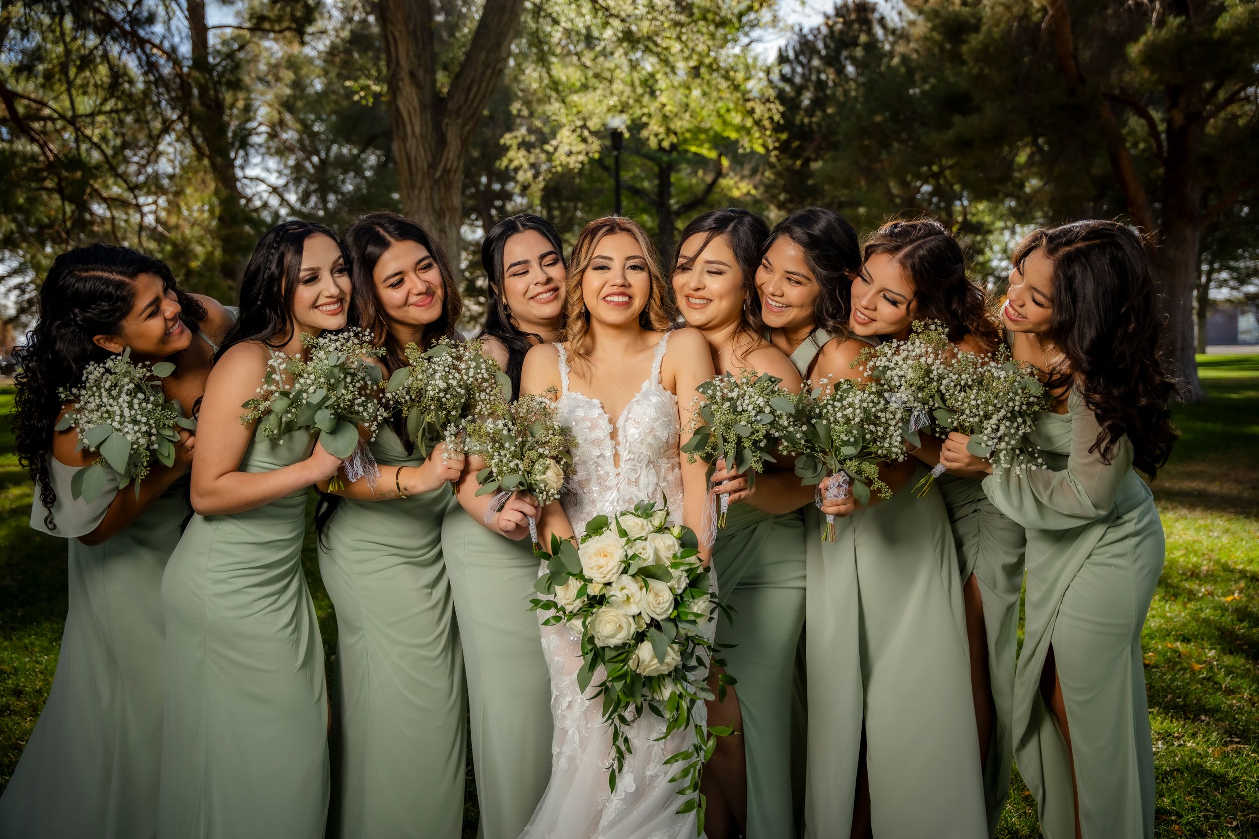 Bride surrounded by bridesmaids in sage green dresses holding white floral bouquets in sunny park.