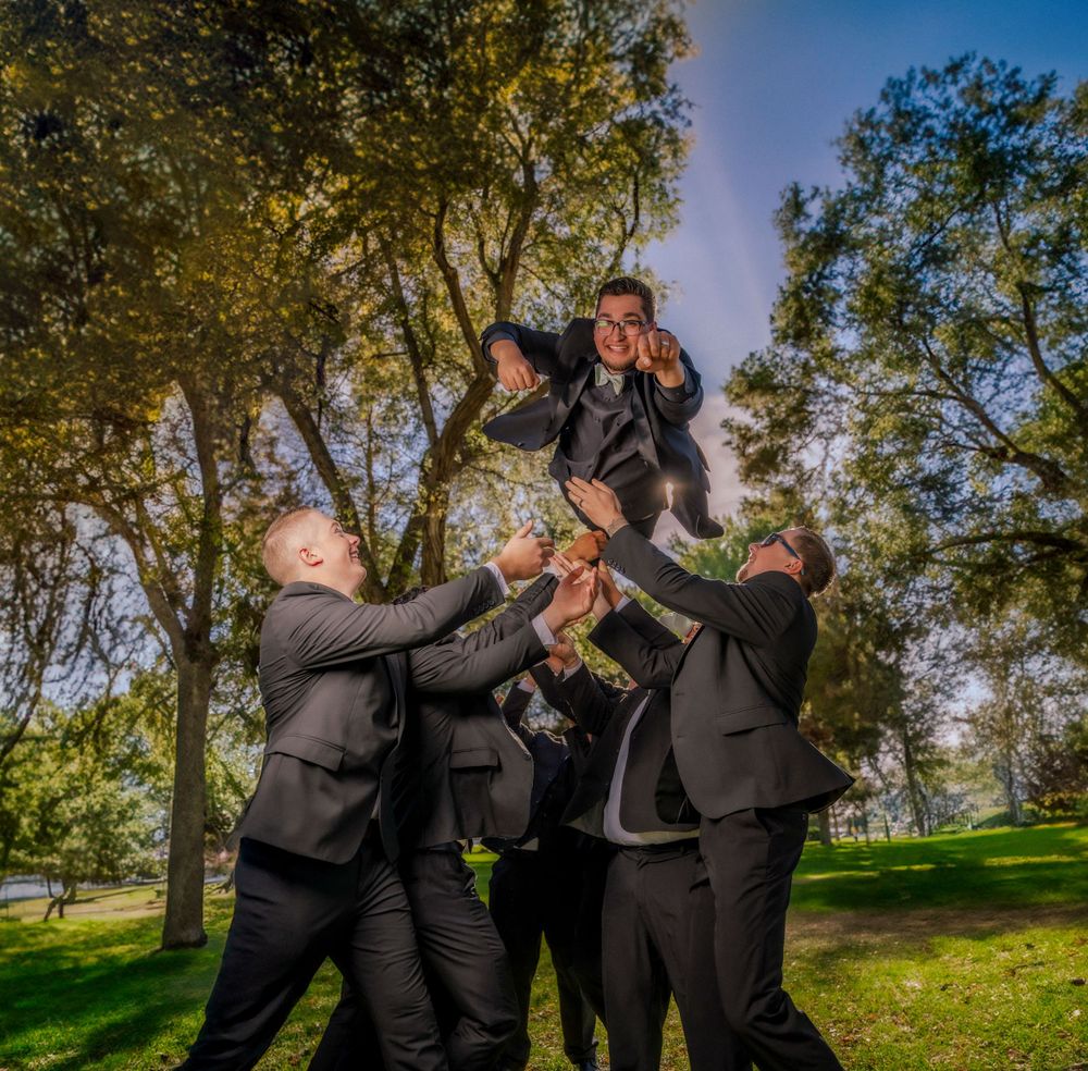 Groomsmen in black suits playfully lift groom in outdoor park wedding party celebration photo.