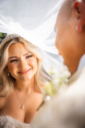 Smiling blonde bride wearing a tiara and veil gazes lovingly at groom during outdoor wedding ceremony.