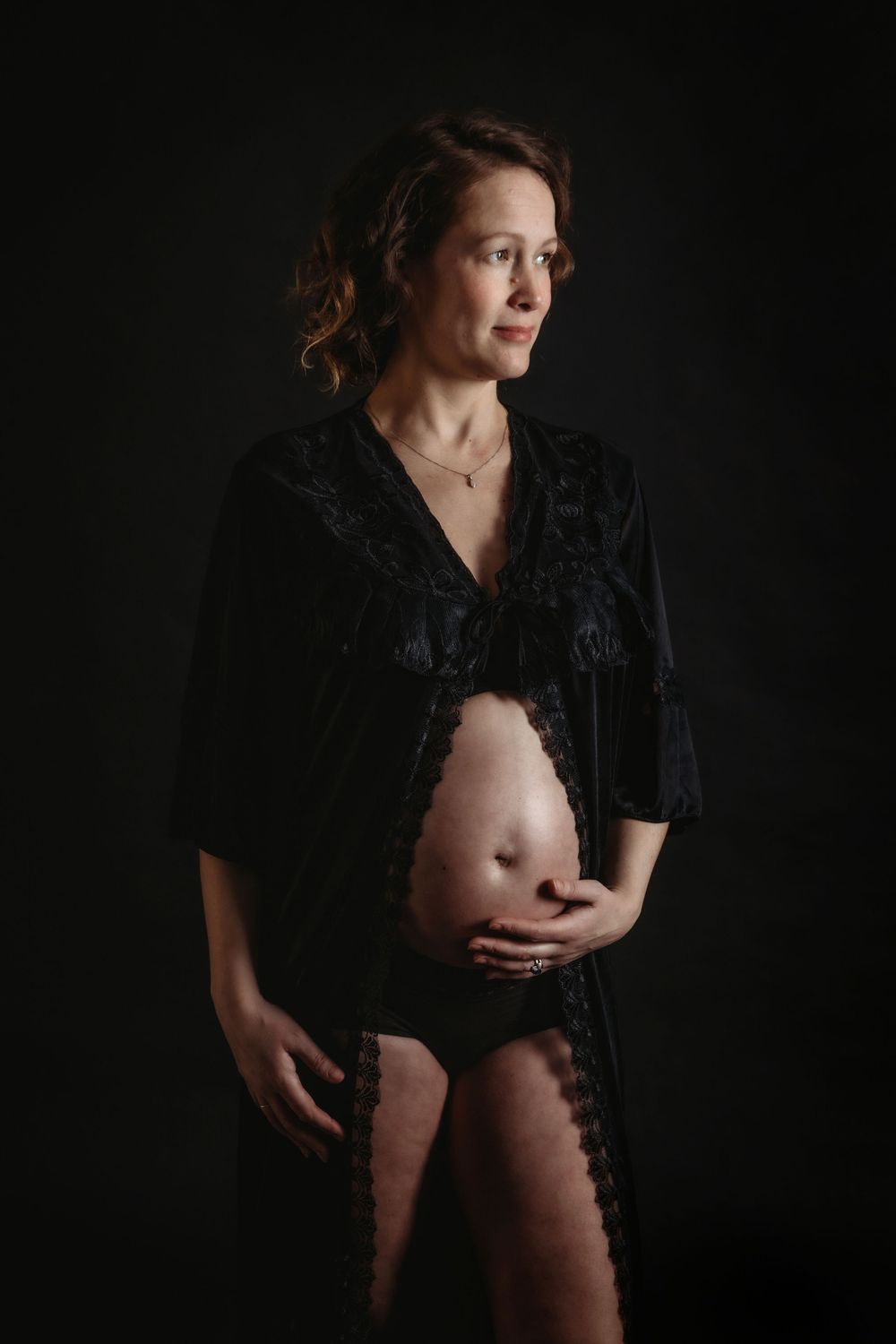 Artistic maternity boudoir photo with black lace robe in dark studio setting.