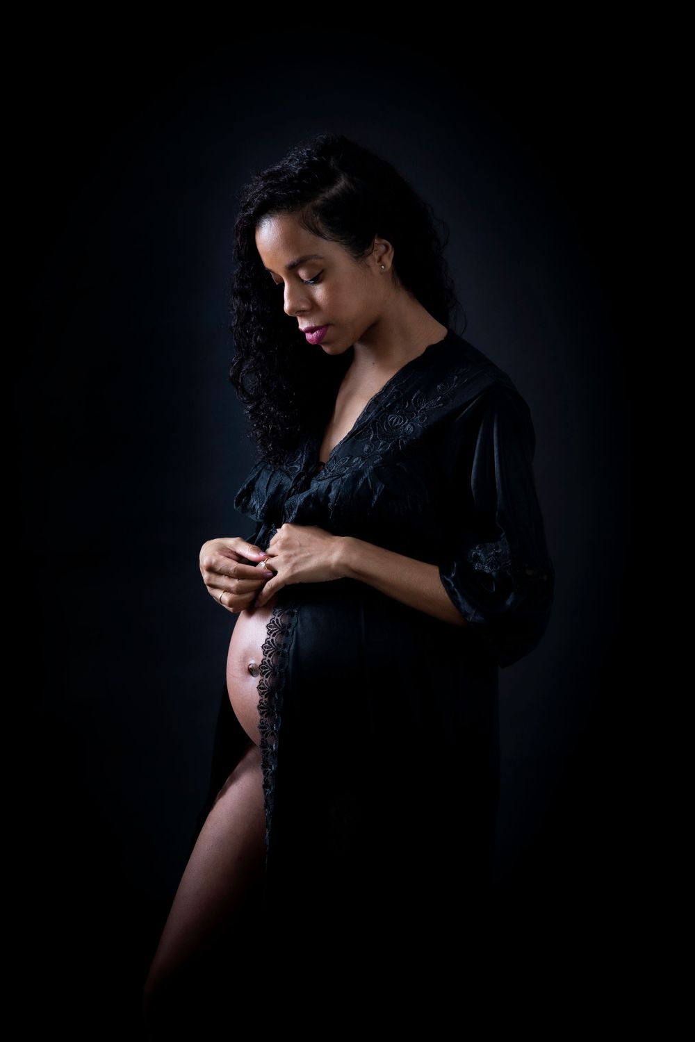 Dramatic maternity portrait in black velvet robe against dark backdrop.