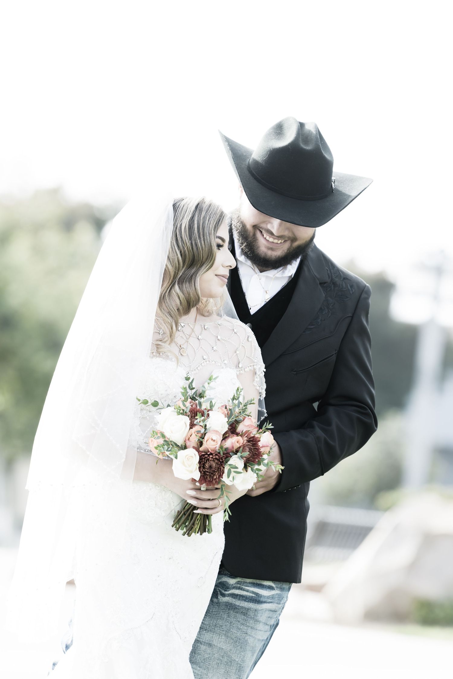 A romantic western-inspired wedding photo with a bride in a lace dress and groom in cowboy hat holding a blush bouquet.