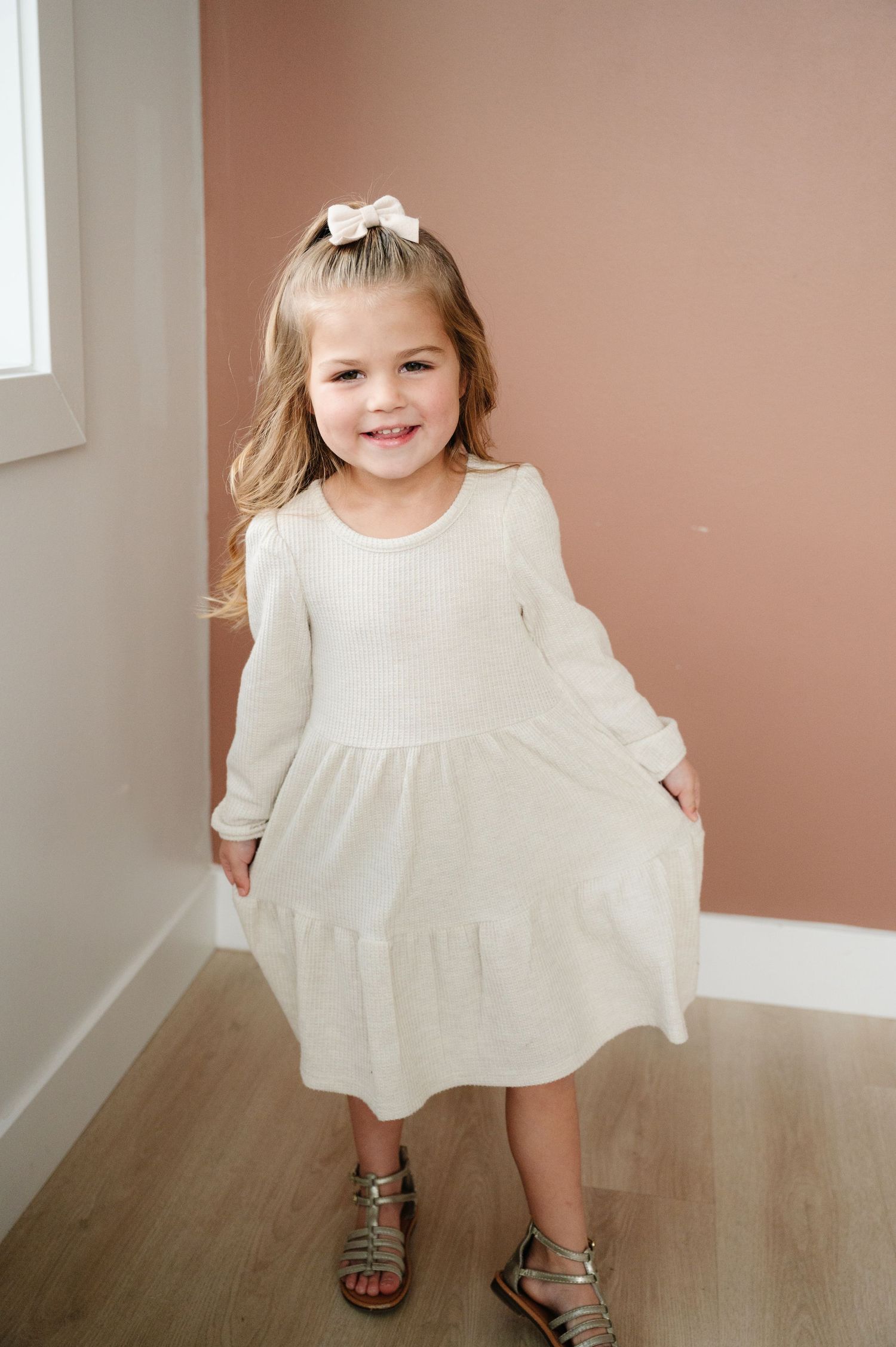 Young child in white dress twirling while standing against pink wall.