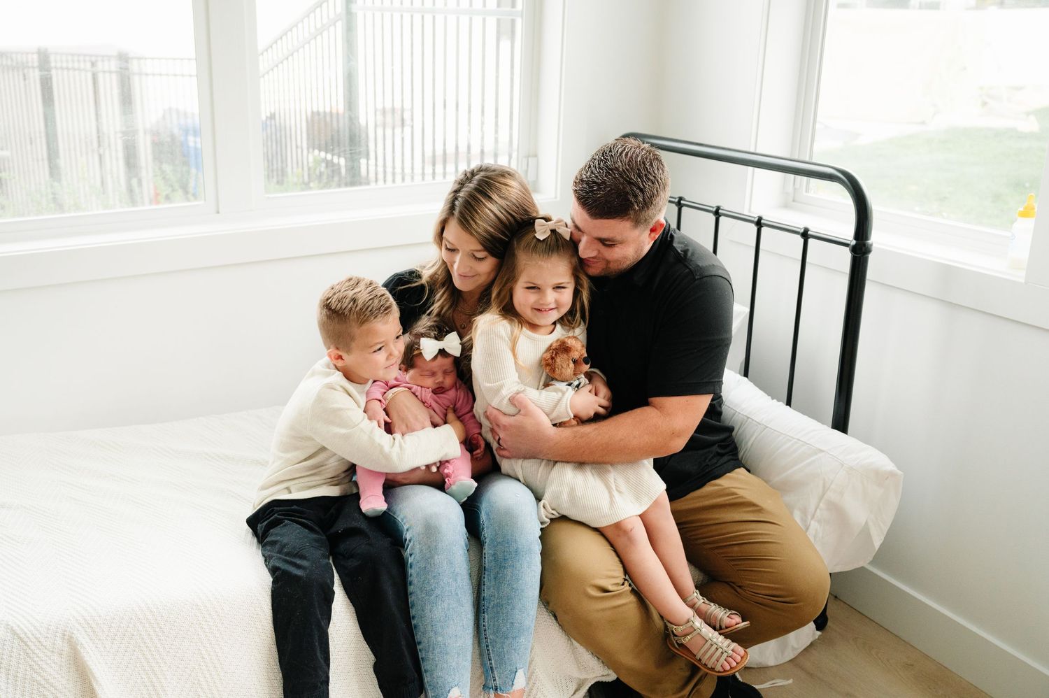 Family cuddles together on a cozy bed with white linens near bright windows.