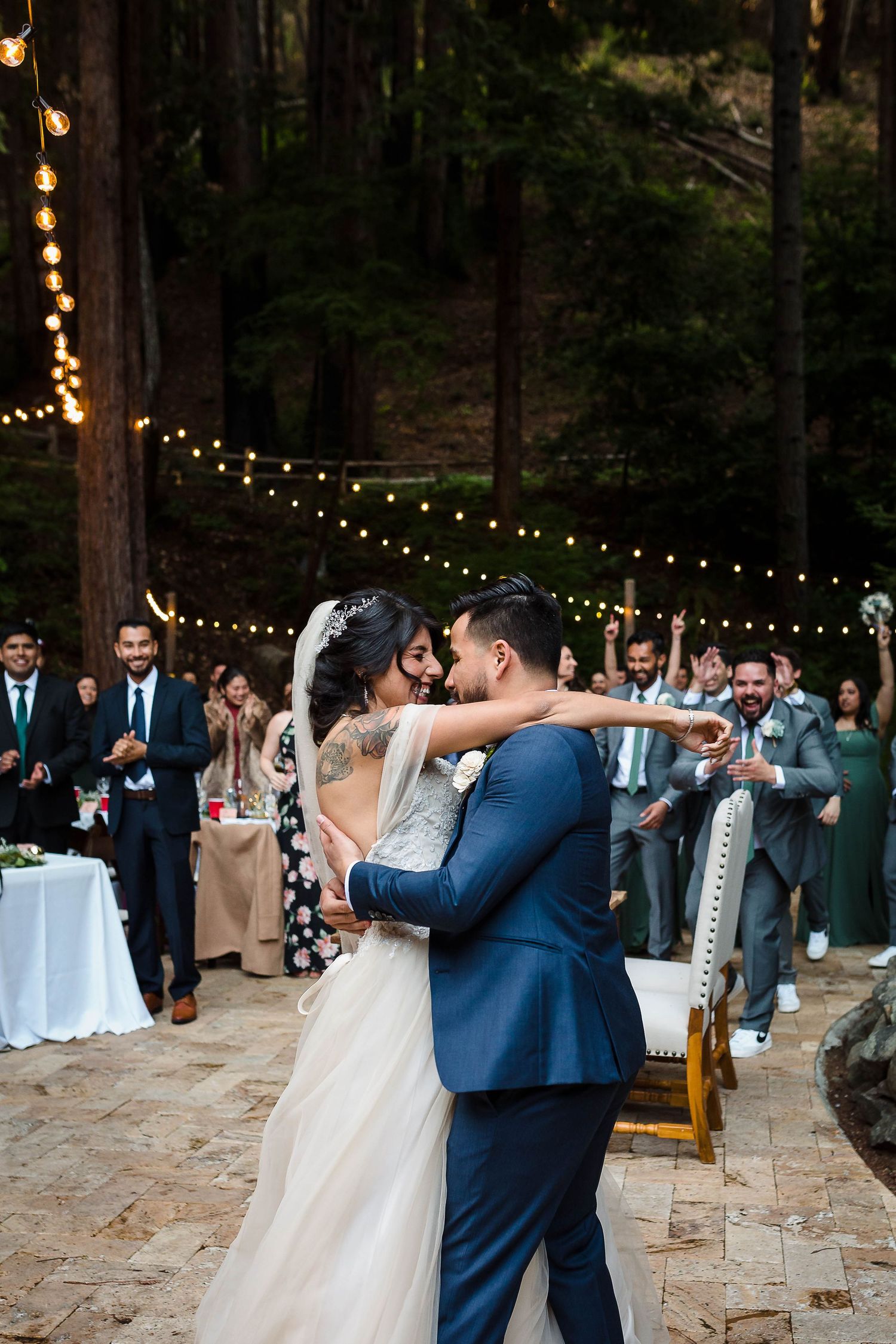 Elegant wedding celebration on stone patio with string lights and redwood trees in background.