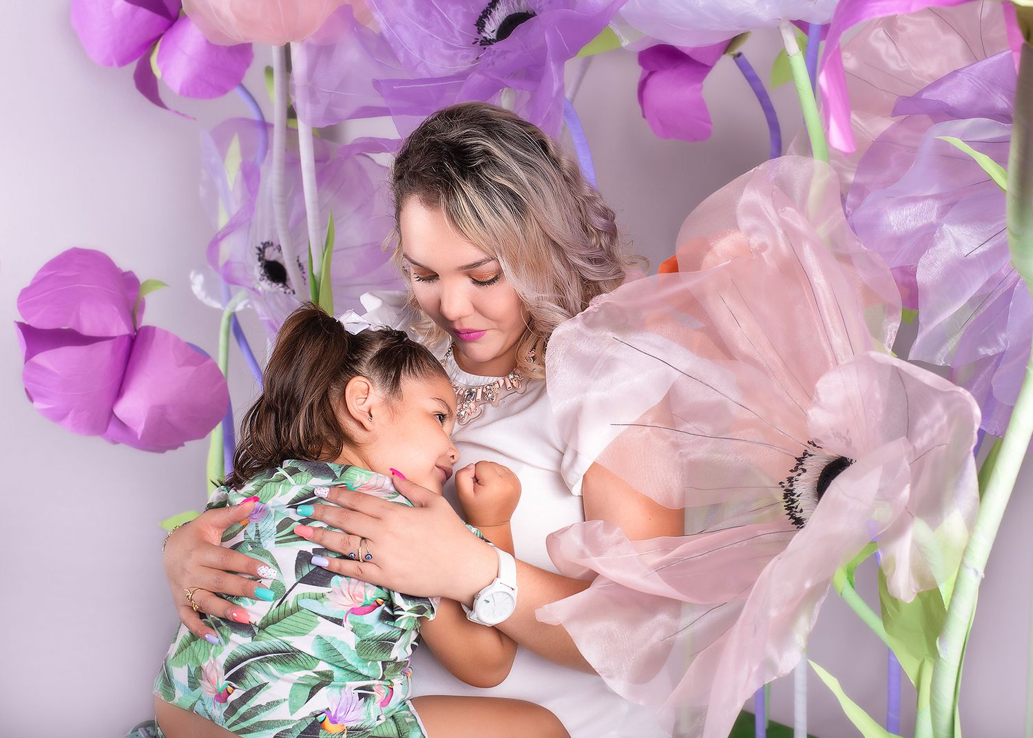 A mother tenderly embraces her two children surrounded by large decorative purple and pink paper flowers.