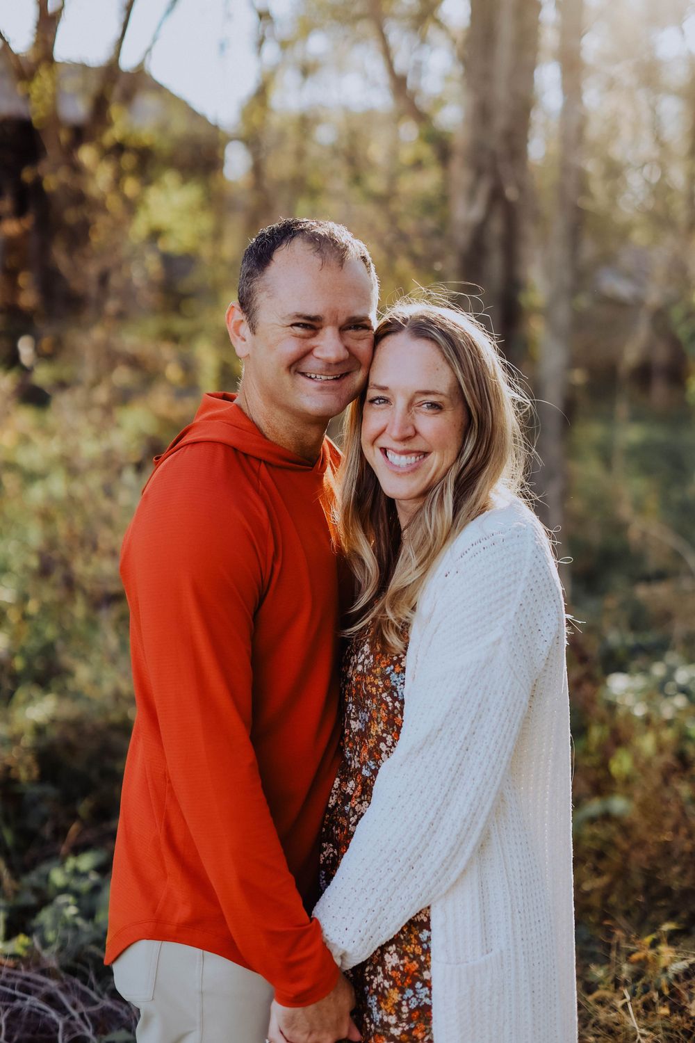 A couple embraces while wearing red and white sweaters in a wooded setting.