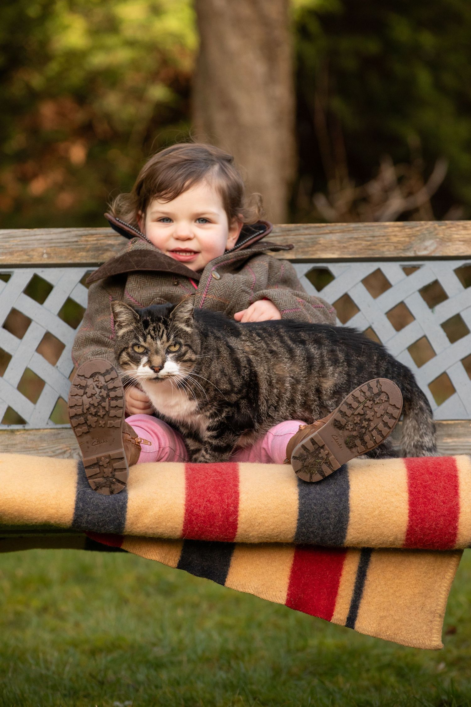 A small child and tabby cat relax together on a colorful striped blanket outdoors.