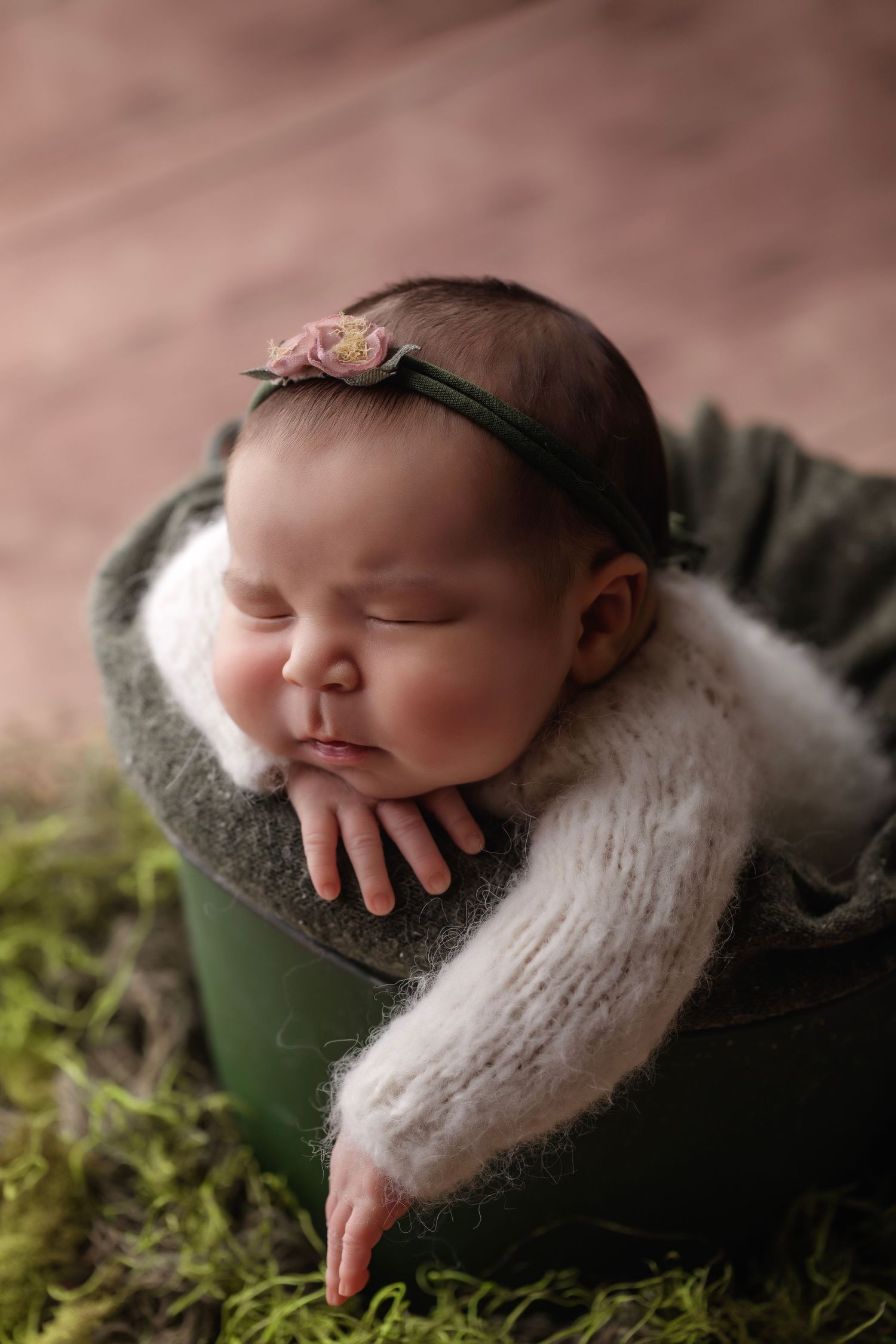 A sleeping newborn with headband rests in a green container surrounded by moss and soft textures.
