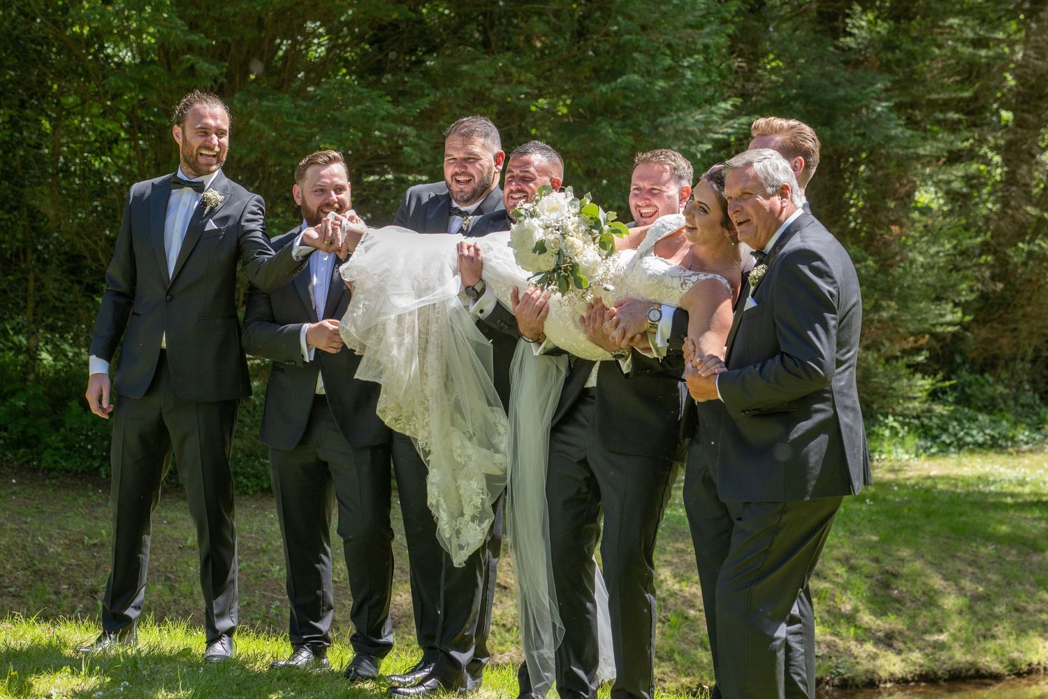 Groomsmen in dark suits playfully lift someone during outdoor wedding photos.