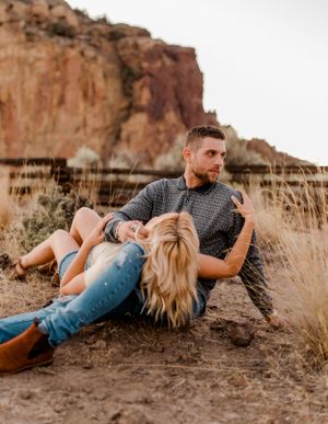 Couple relaxing on desert terrain with dramatic red cliffs in background.