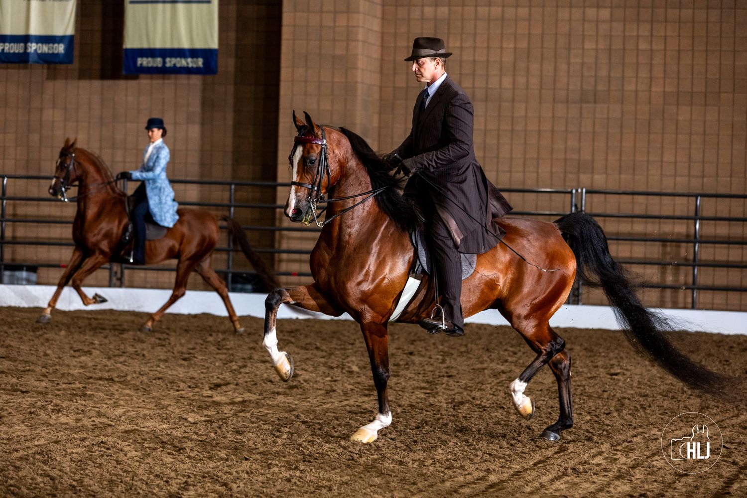 Two riders performing dressage movements on bay horses in an indoor arena setting.