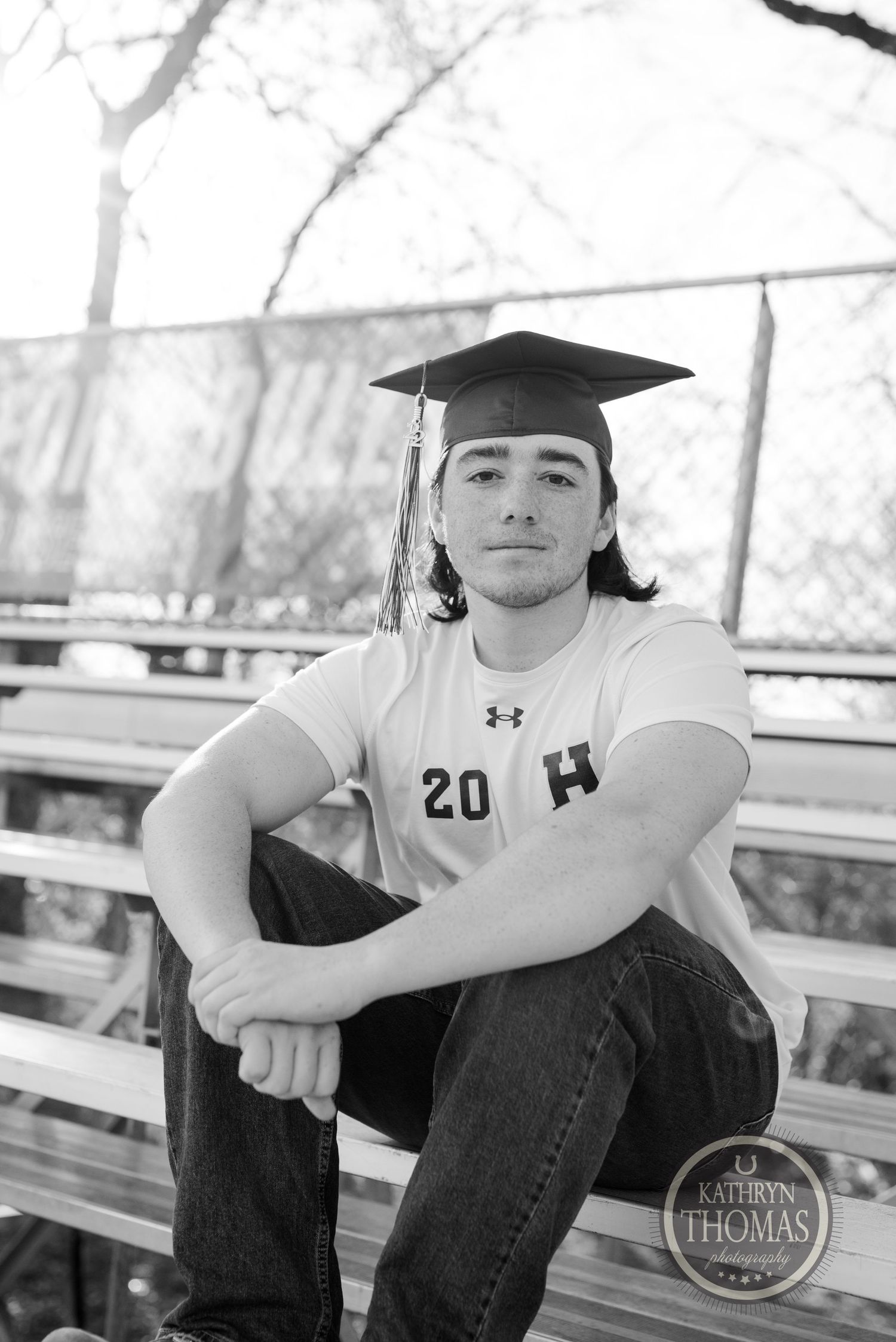 Black and white portrait of graduate in cap sitting on bleachers during senior photoshoot.