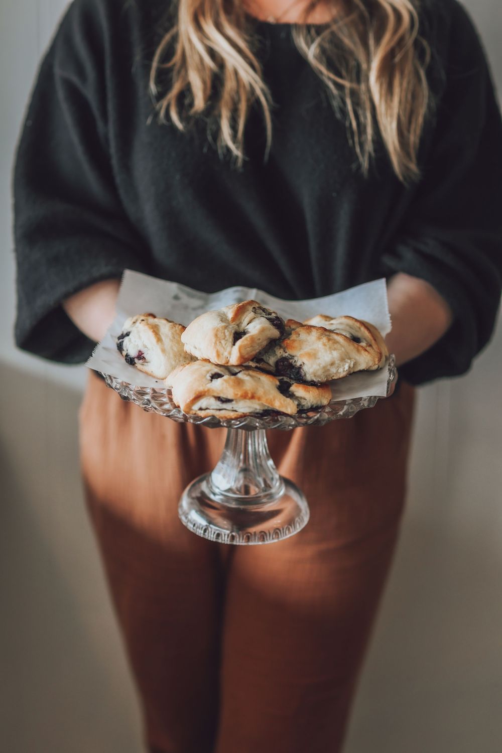 Person holding pastries on decorative cake stand wearing black top and brown pants.