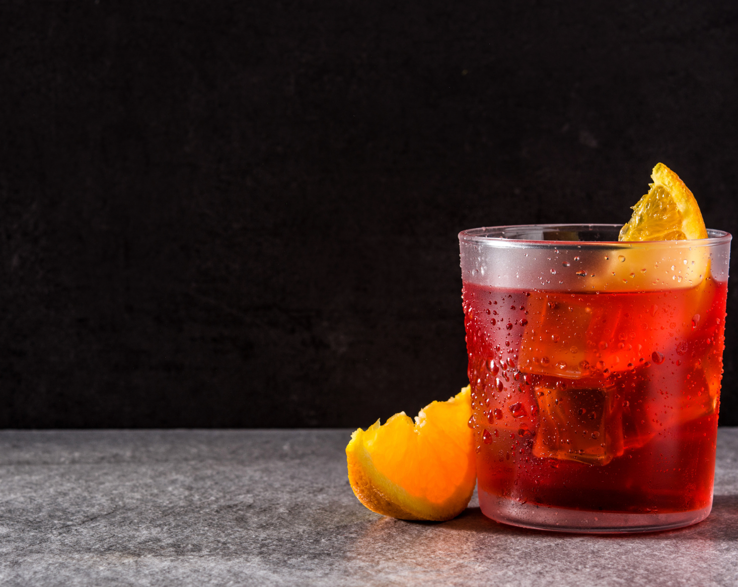 Red cocktail with orange garnish served in rocks glass on dark background.