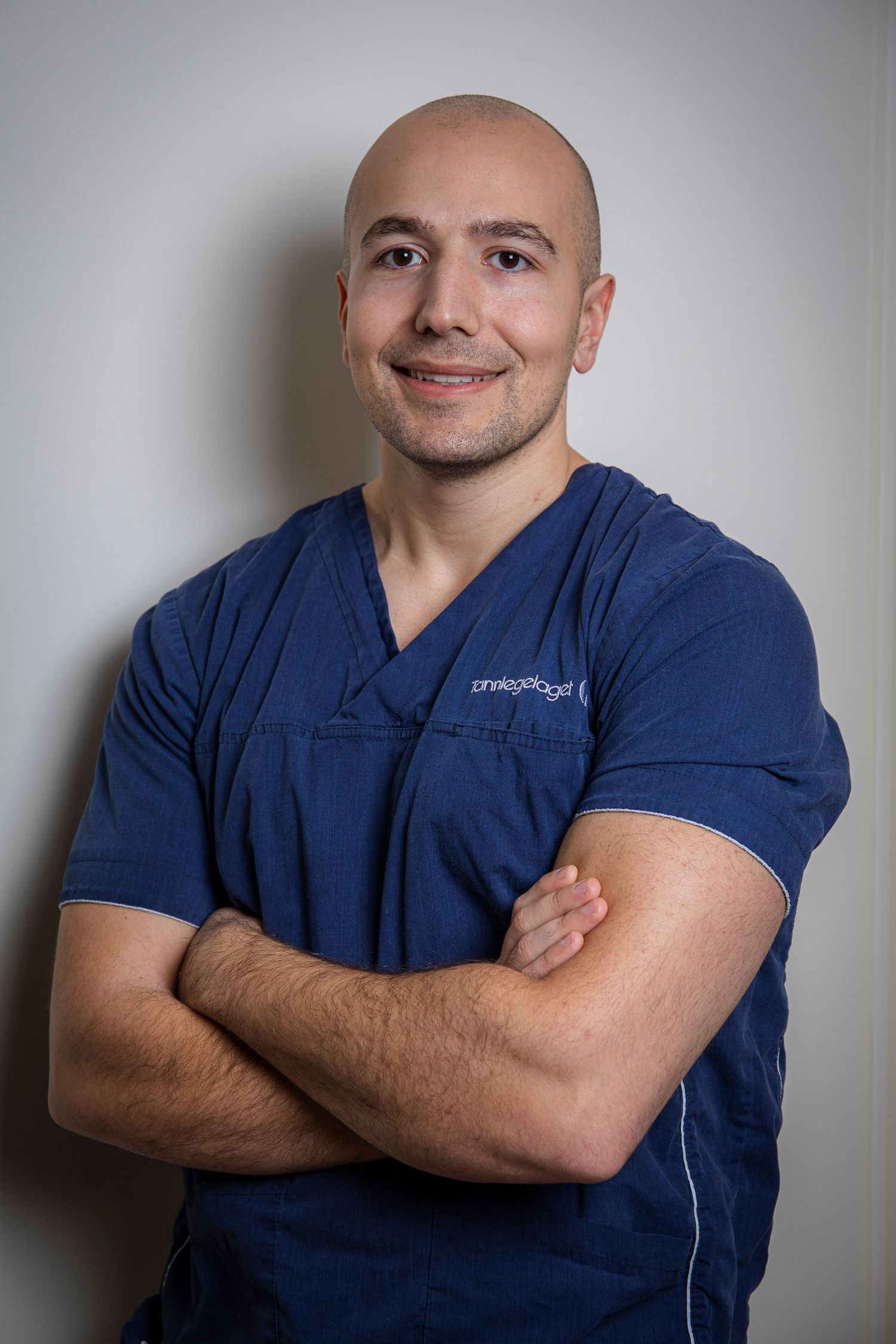 Bald male healthcare professional in navy scrubs with arms crossed smiling confidently.