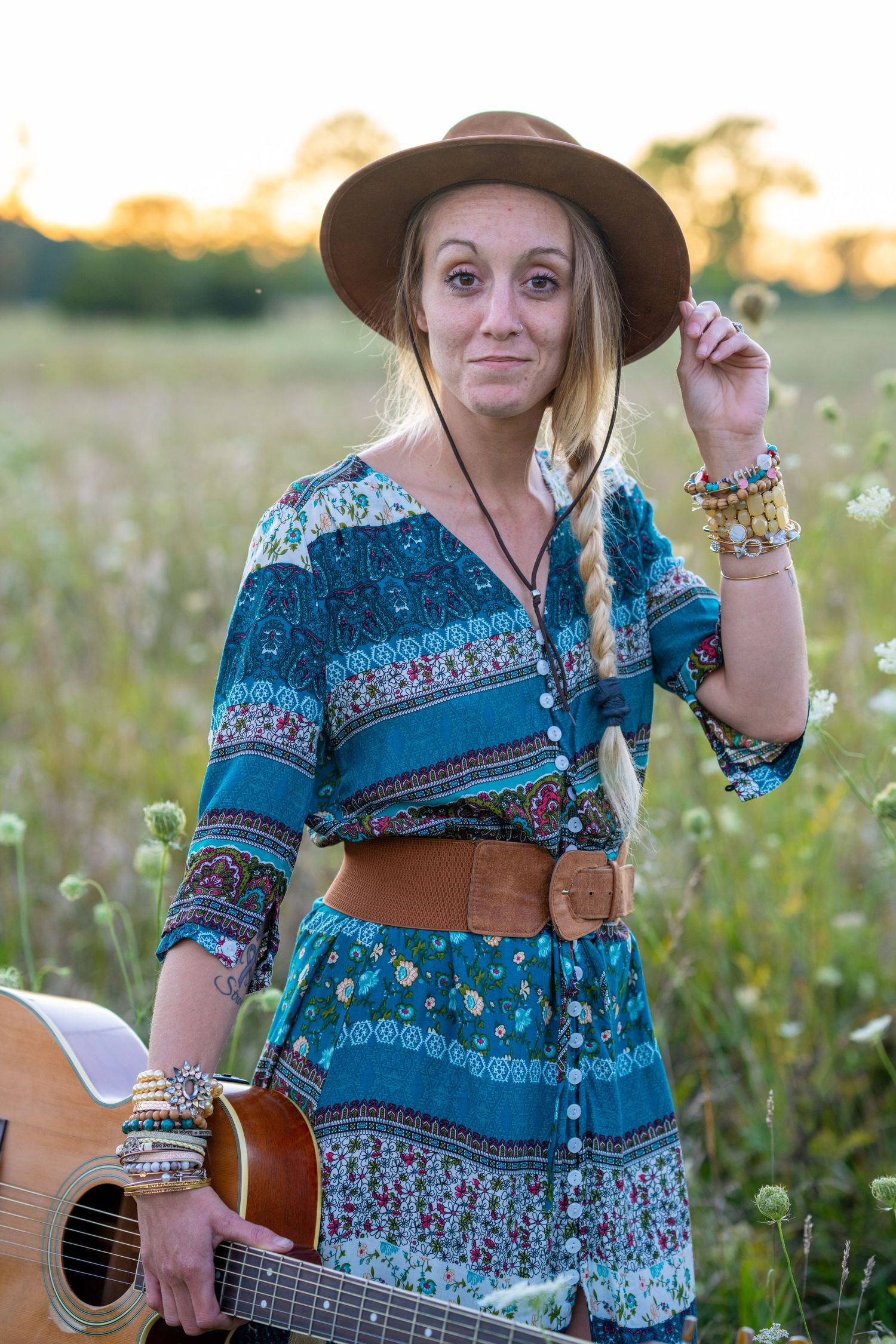 Country musician in blue patterned dress and brown hat poses with guitar in wildflower field.