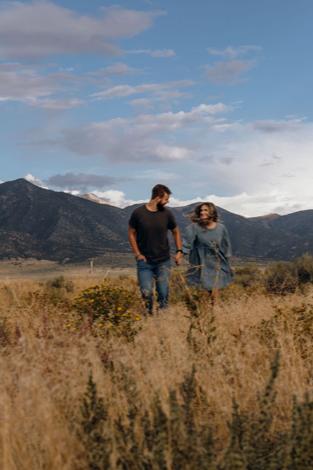 A couple walking together through tall grass with mountains and blue sky in the background.