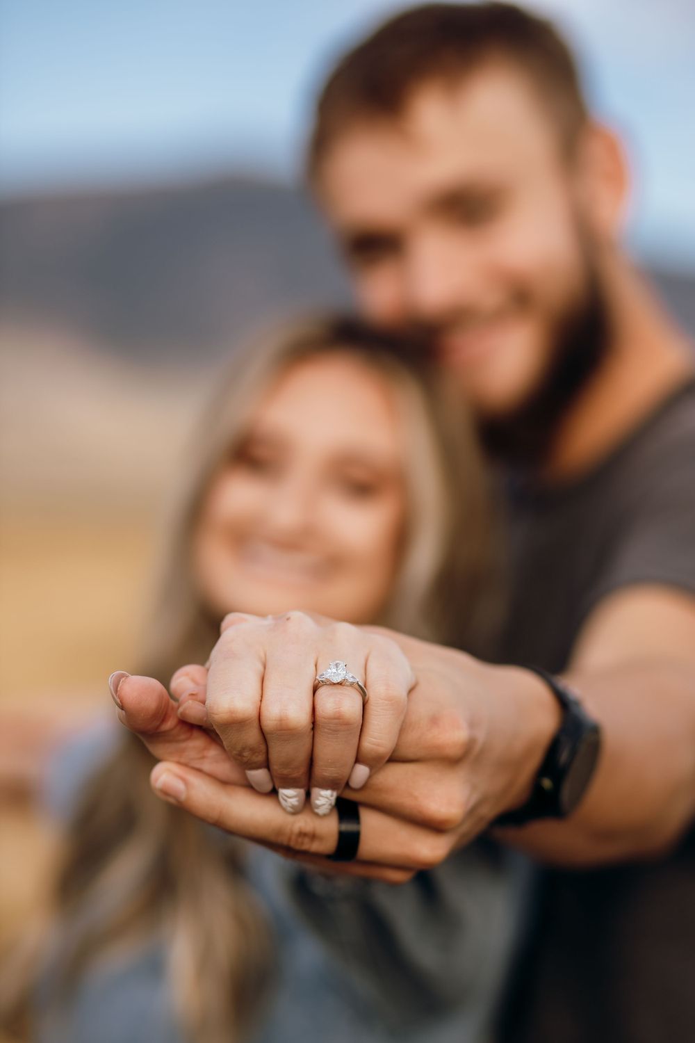 A close-up shot of hands showing off an engagement ring against a mountain backdrop.