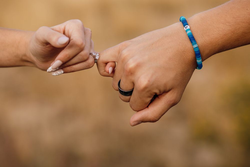 Two hands linking pinky fingers with one wearing a blue bracelet against a warm outdoor background.