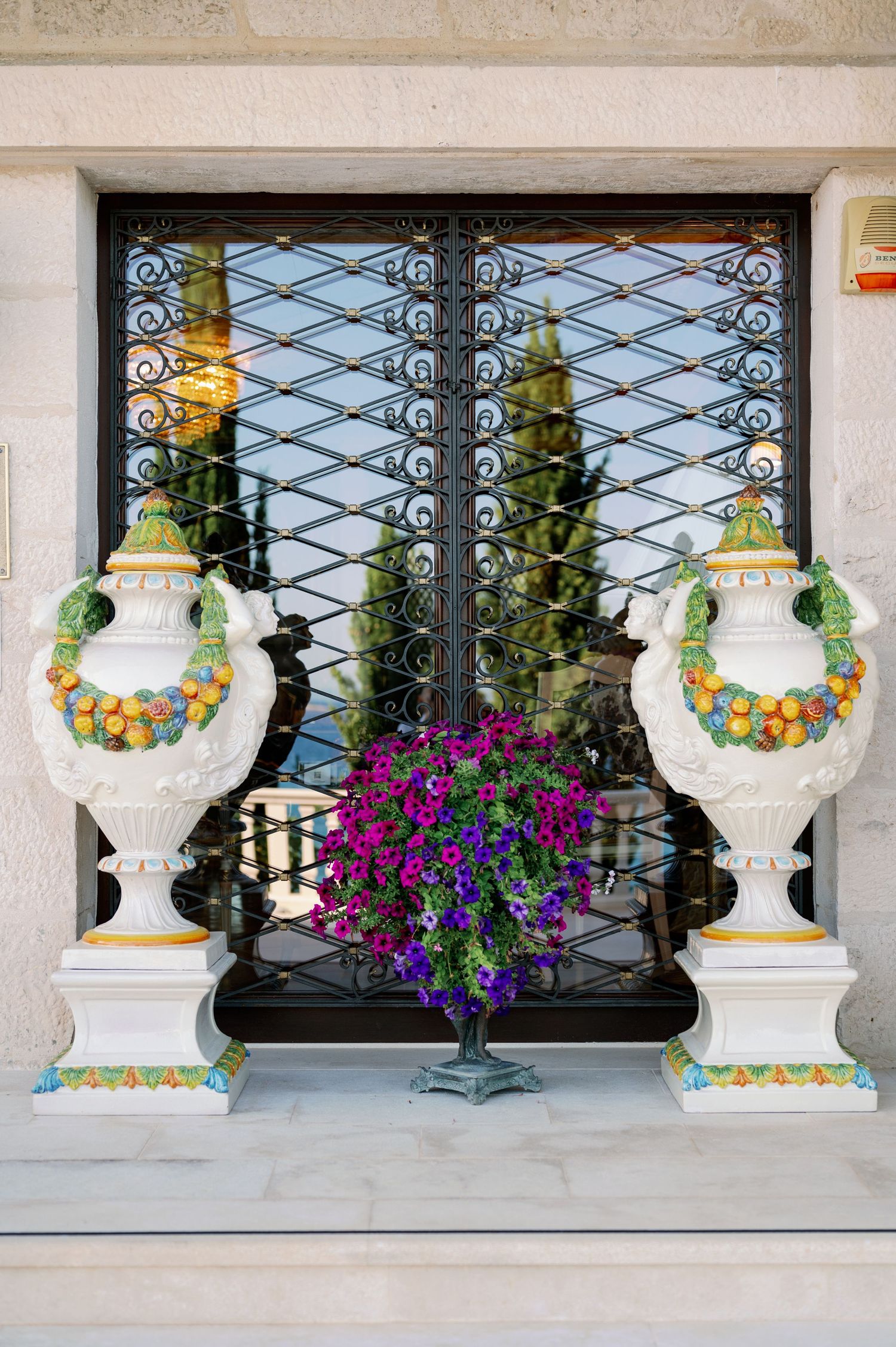 Ornate decorative urns and purple flowers adorn a window with intricate metal grating.
