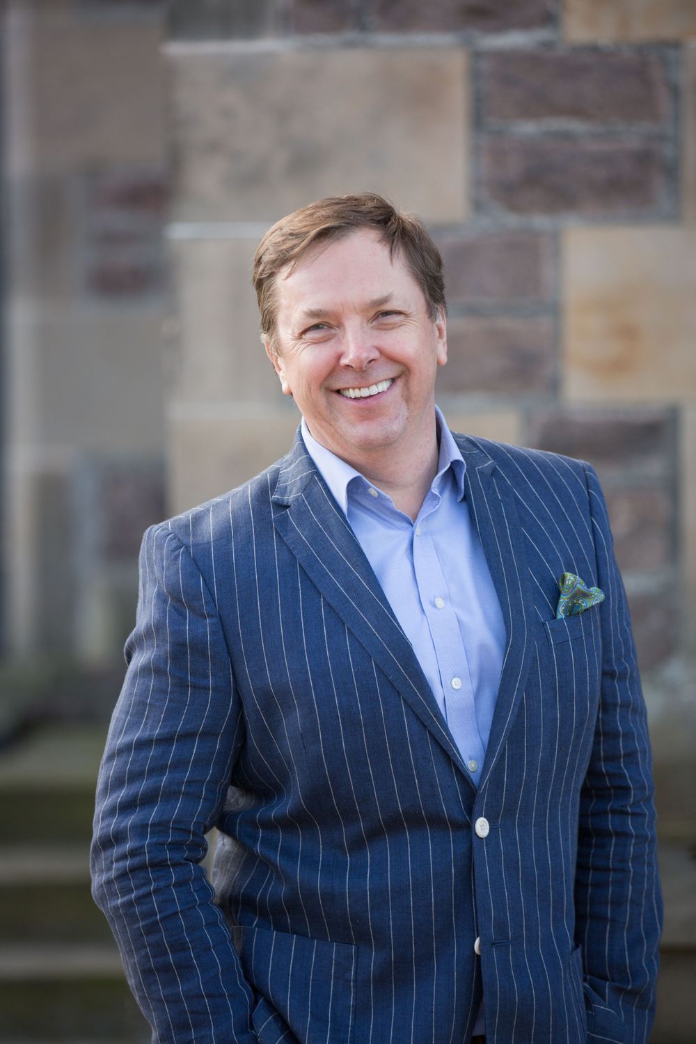 Professional wearing a navy pinstripe suit with light blue shirt and pocket square outdoors.