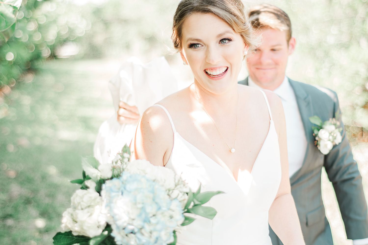 A couple in wedding attire stands close together outdoors holding a light blue and white floral bouquet in sunlit setting.
