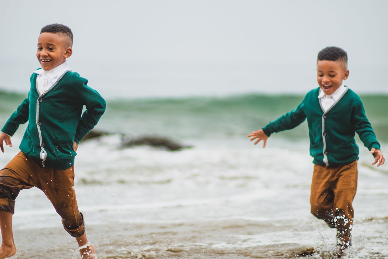 Two children in matching green sweaters run and play at the beach.