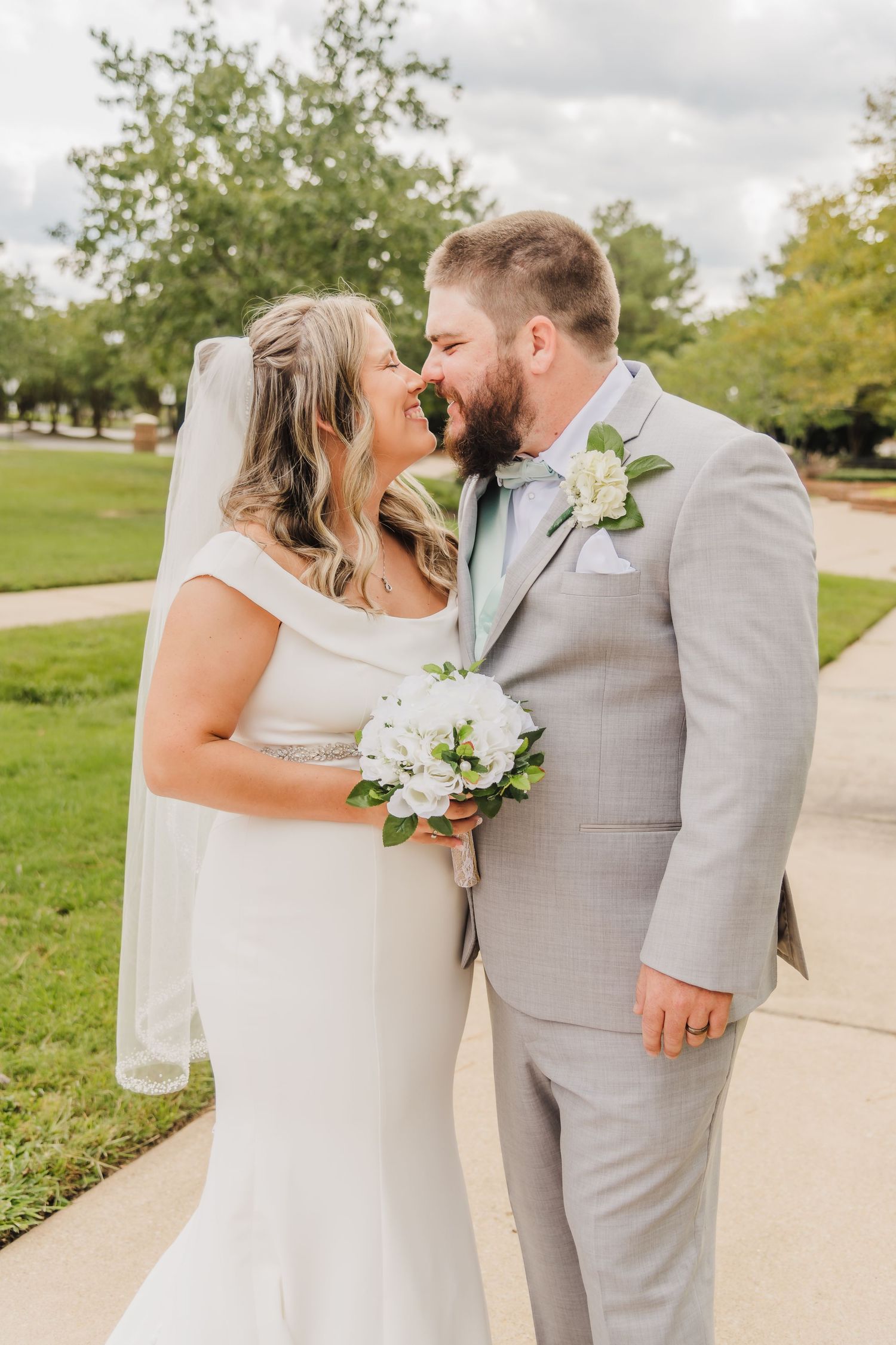 Newlywed couple in light gray suit and white dress share an intimate moment with white bouquet on their wedding day.