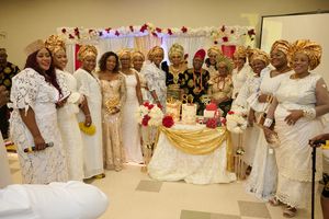 Group photo of guests in elegant white and gold attire gathered around a decorated cake table.
