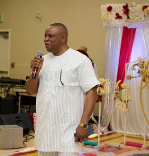 Speaker in white traditional Nigerian attire addresses audience at decorated event venue.